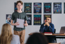 Picture shows a young boy standing up and speaking in class as the teacher watches with pride.