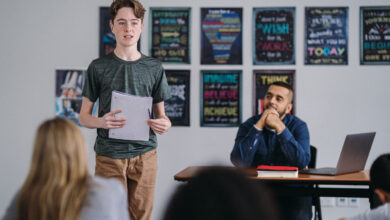 Picture shows a young boy standing up and speaking in class as the teacher watches with pride.
