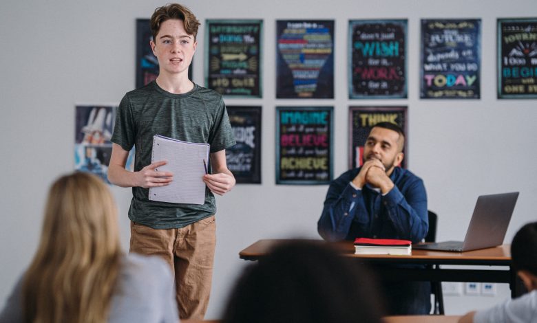 Picture shows a young boy standing up and speaking in class as the teacher watches with pride.