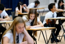 Teenage girl wearing school shirt and tie writing in exam booklet, arm resting on desk