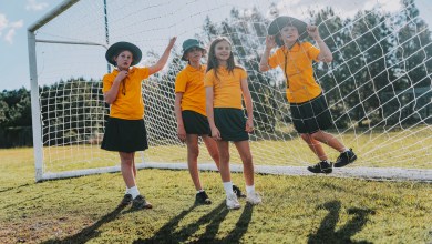 Four schoolgirls in yellow polo shirts and sun hats lean and play against a soccer goal net on a grassy sports field in Byron Bay, one resting a foot on the frame during a relaxed break from school sport.