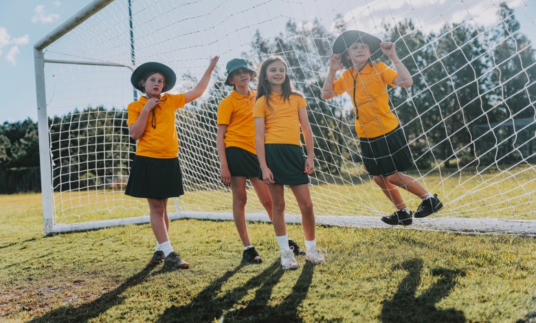 Four schoolgirls in yellow polo shirts and sun hats lean and play against a soccer goal net on a grassy sports field in Byron Bay, one resting a foot on the frame during a relaxed break from school sport.